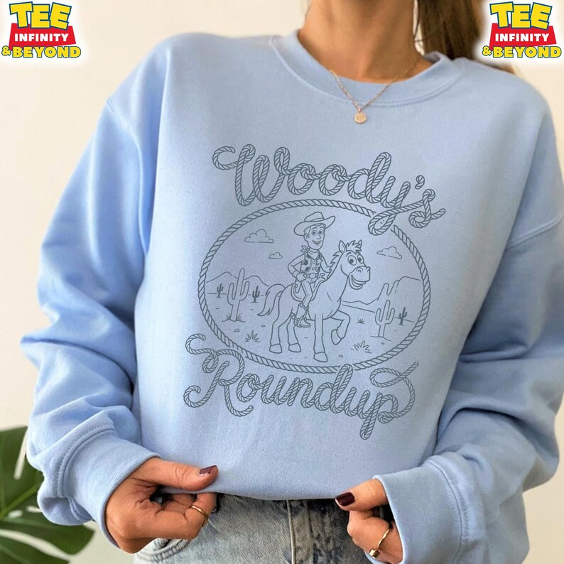 May include: A light blue crewneck sweatshirt featuring a dark blue line art graphic of a cowboy riding a horse within a rope-style oval frame. The text Woody's Roundup is written in a rope-style font above and below the central illustration.