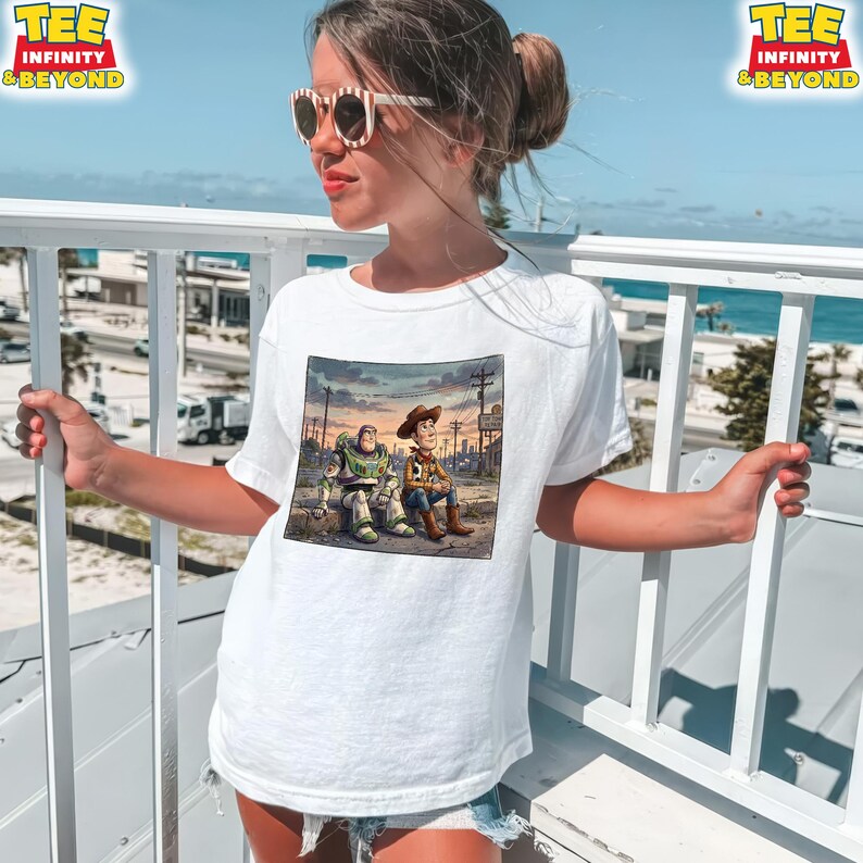 May include: White t-shirt with a colourful illustration of Buzz Lightyear and Woody from Toy Story. The graphic shows the characters sitting together in a sunset scene. The shirt has the text "TEE INFINITY & BEYOND" in the upper left corner.