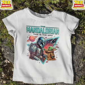 Może przedstawiać: Biały t-shirt z nadrukiem graficznym przedstawiającym Mandalorianina i Grogu. Tekst głosi THE MANDALORIAN THIS IS THE WAY. Projekt zawiera planetę, gwiazdy i tr&oacute;jkątny element tła w kolorowym, ilustrowanym stylu komiksowym.