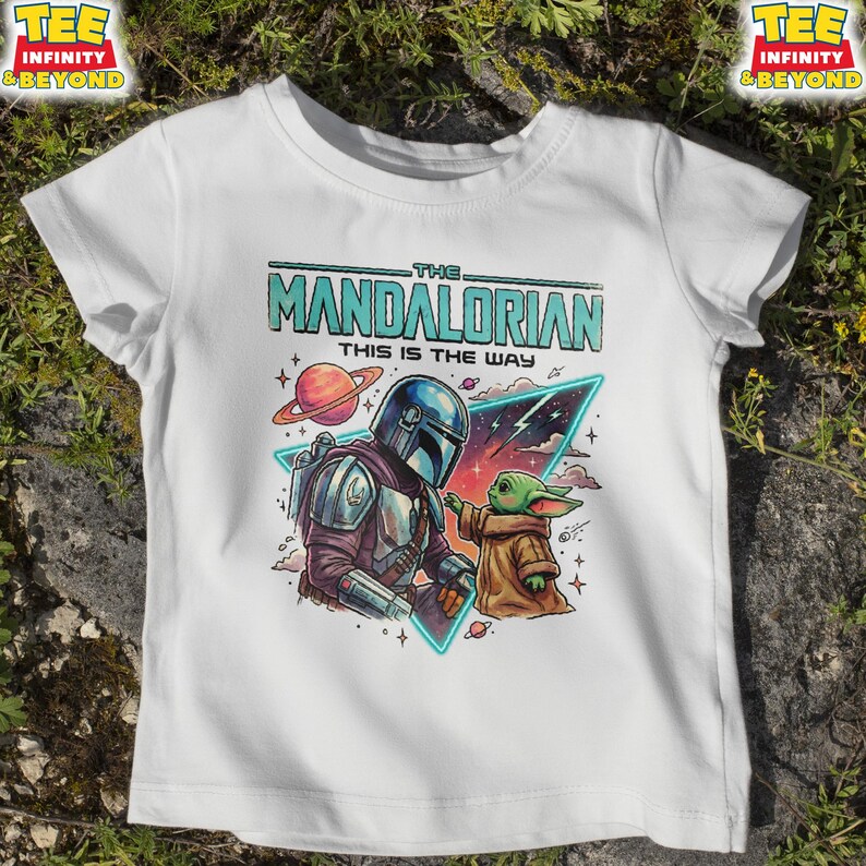 Może przedstawiać: Biały t-shirt z nadrukiem graficznym przedstawiającym Mandalorianina i Grogu. Tekst głosi THE MANDALORIAN THIS IS THE WAY. Projekt zawiera planetę, gwiazdy i tr&oacute;jkątny element tła w kolorowym, ilustrowanym stylu komiksowym.