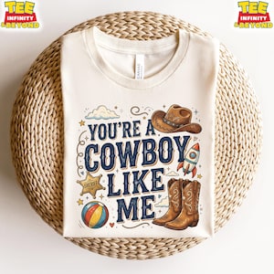 May include: A cream-colored t-shirt featuring the text "YOU'RE A COWBOY LIKE ME" in dark blue lettering. The design includes illustrations of a brown cowboy hat, a sheriff star badge, a colorful beach ball, a space rocket, and brown cowboy boots.