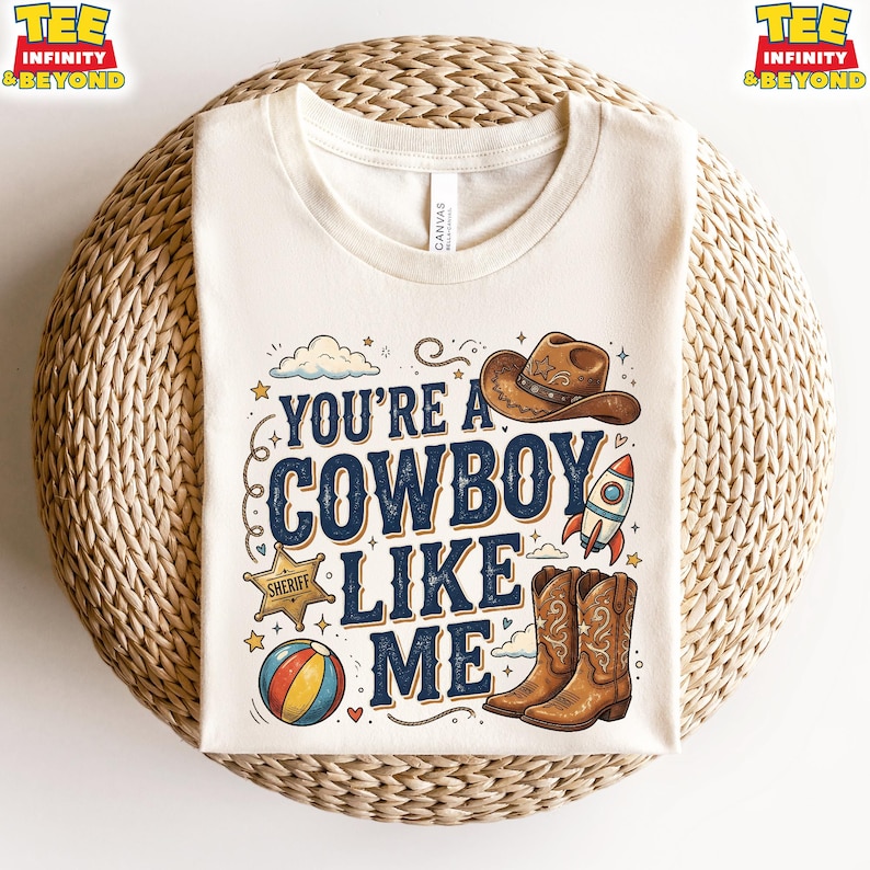 May include: A cream-colored t-shirt featuring the text "YOU'RE A COWBOY LIKE ME" in dark blue lettering. The design includes illustrations of a brown cowboy hat, a sheriff star badge, a colorful beach ball, a space rocket, and brown cowboy boots.