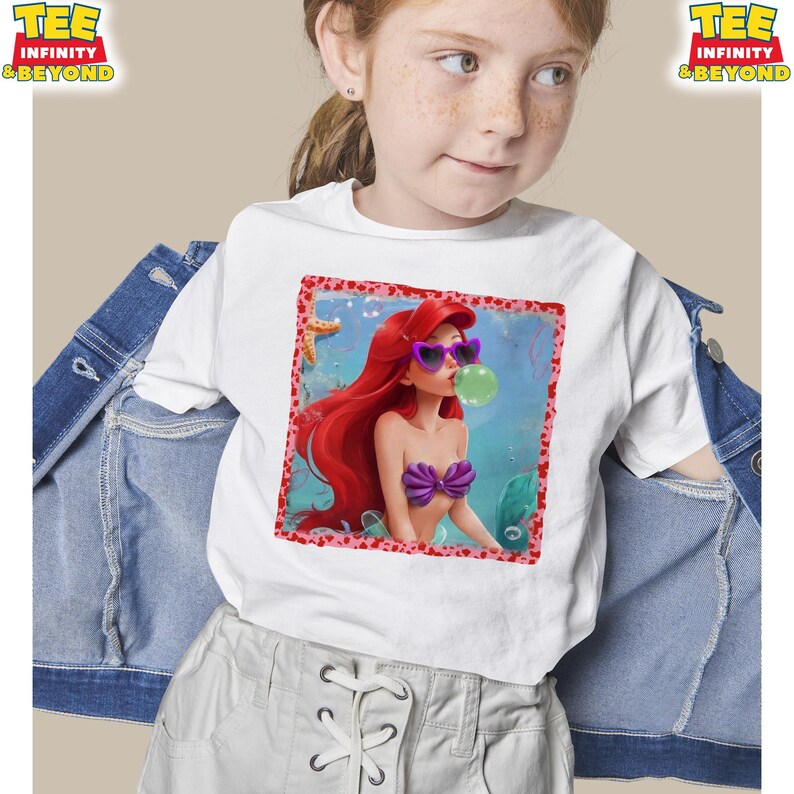 May include: White t-shirt featuring a colorful graphic of a cartoon mermaid with red hair, purple sunglasses, and a purple seashell top blowing a bubble. The image is framed with a red border. The shirt has the text "TEE INFINITY & BEYOND" on the top.