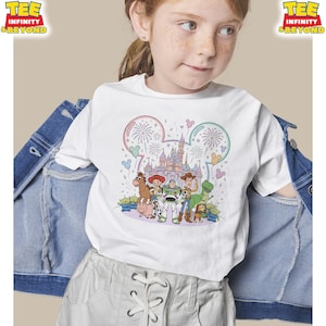 May include: White t-shirt featuring a colorful graphic of Disney's Toy Story characters, including Woody, Buzz Lightyear, and Rex, with a castle and fireworks design. The shirt has the text "TEE INFINITY & BEYOND" at the top.