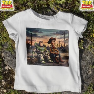 May include: White t-shirt with a graphic of Buzz Lightyear and Woody from Toy Story, set against a sunset. The design includes the text "TEE INFINITY & BEYOND" in a yellow and red logo. The image depicts the characters sitting together.