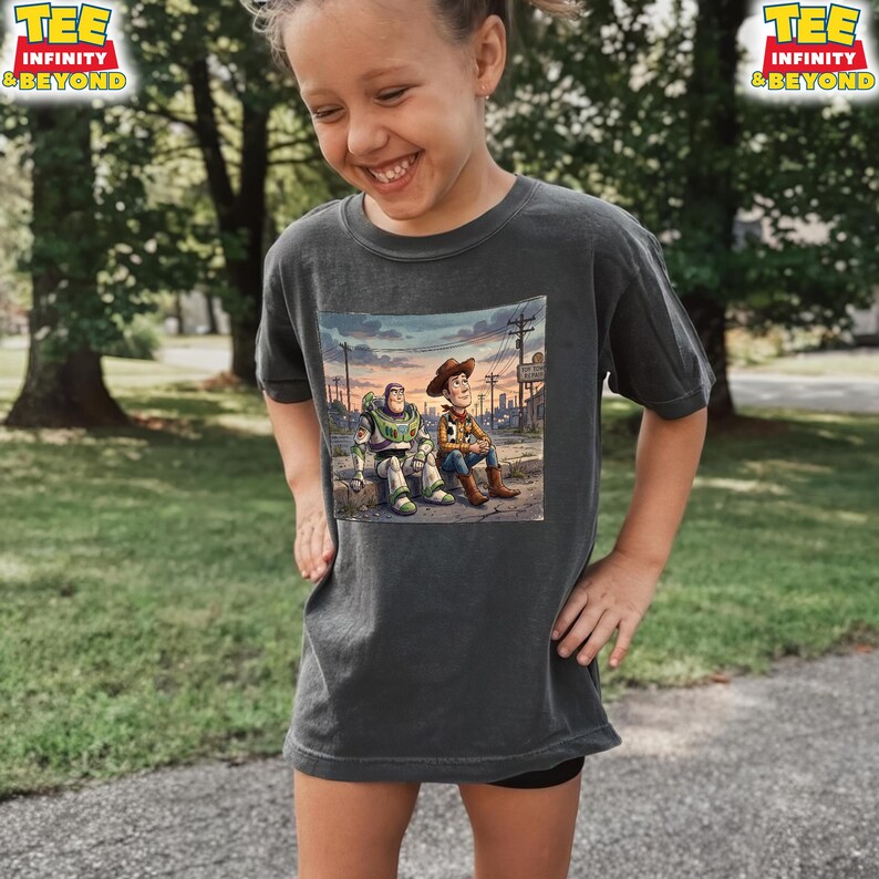 May include: A dark grey t-shirt with a colourful graphic of Buzz Lightyear and Woody from Toy Story. The image shows the characters together in a sunset scene. The shirt has the text "TEE INFINITY & BEYOND" in a yellow and red logo.