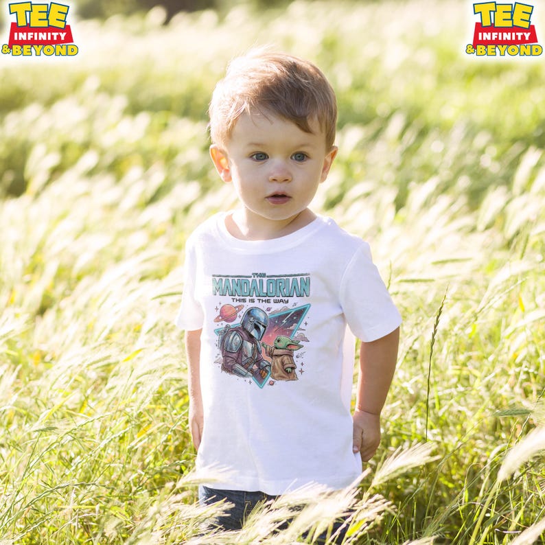 Może przedstawiać: Biały t-shirt z kr&oacute;tkim rękawem z nadrukiem graficznym przedstawiającym Mandalorianina i Grogu. Wz&oacute;r zawiera napis "THE MANDALORIAN" oraz "THIS IS THE WAY" na kolorowym tle kosmicznym z planetą i gwiazdami.