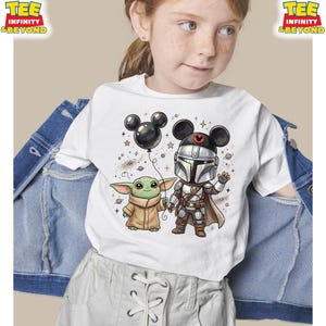 May include: A white short-sleeved t-shirt featuring a graphic illustration of a character in silver armor with a black Mickey Mouse ear hat and a small green creature in a tan robe holding a black balloon, surrounded by stars and planets.