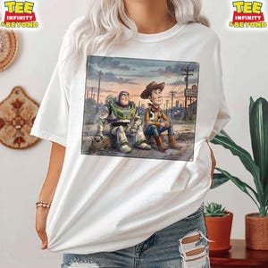 May include: White t-shirt with a colourful illustration of Buzz Lightyear and Woody from Toy Story. The graphic shows them sitting together against a cityscape backdrop with a "Toy Town Repair" sign. The shirt has the text "TEE INFINITY & BEYOND" on the top left and right.