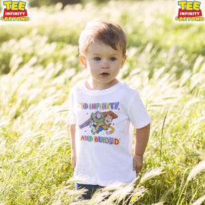 May include: A white t-shirt with a colourful graphic of Buzz Lightyear and Woody from Toy Story, and the text "TO INFINITY AND BEYOND." A young child wears the shirt while standing in a field of tall grass. The shirt has the words "TEE INFINITY & BEYOND" on the top left.