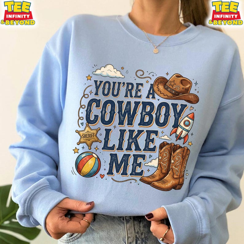 May include: A light blue crewneck sweatshirt featuring a graphic print with the text "YOU'RE A COWBOY LIKE ME". The design includes a brown cowboy hat, a gold sheriff star badge, a colorful beach ball, a rocket ship, and brown cowboy boots.
