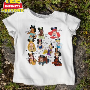 May include: White t-shirt with a graphic of Disney princesses wearing mouse ears. The princesses include Jasmine, Tiana, Ariel, Snow White, Cinderella, Aurora, Belle, Mulan, and  Mowgli. Each princess has her name written next to her.