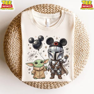 May include: A cream-colored t-shirt featuring a graphic of a small green creature in a tan robe and a silver-armored character wearing a black Mickey Mouse ear hat. The graphic includes a black balloon, stars, and planets in a cartoon style.