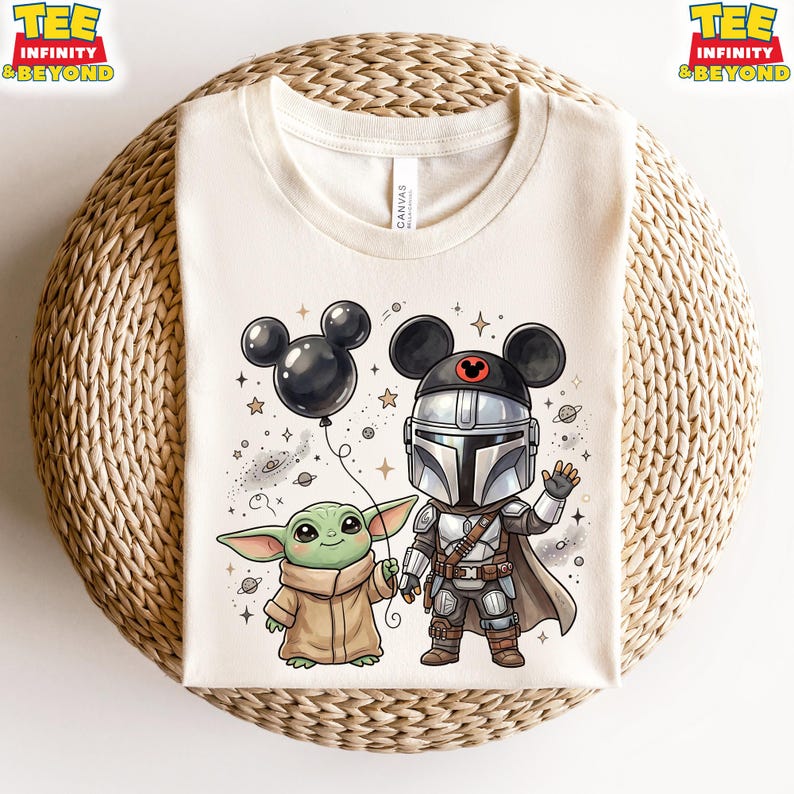 May include: A cream-colored t-shirt featuring a graphic of a small green creature in a tan robe and a silver-armored character wearing a black Mickey Mouse ear hat. The graphic includes a black balloon, stars, and planets in a cartoon style.