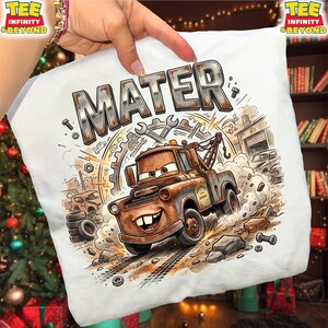 May include: White t-shirt featuring a cartoon illustration of Mater from the movie Cars. The design includes the name "MATER" in large, metallic letters, along with tools, gears, and a garage scene. The image is a detailed, colorful graphic.