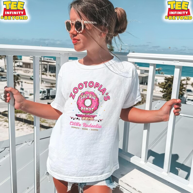 Puede incluir: Camiseta blanca con un gr&aacute;fico de donut rosa y el texto "Zootopia's The Big Donut" y "Little Rodentia Donuts - Bagel - Scoffee". La camiseta tiene mangas cortas.