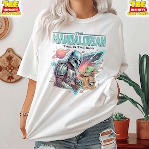 Może przedstawiać: Biały t-shirt z nadrukiem graficznym przedstawiającym Mandalorianina i Grogu. Wz&oacute;r zawiera napis "THE MANDALORIAN THIS IS THE WAY" nad postaciami, na tle kolorowej przestrzeni kosmicznej z planetą i neonowym tr&oacute;jkątem.