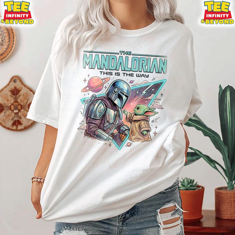 Może przedstawiać: Biały t-shirt z nadrukiem graficznym przedstawiającym Mandalorianina i Grogu. Wz&oacute;r zawiera napis "THE MANDALORIAN THIS IS THE WAY" nad postaciami, na tle kolorowej przestrzeni kosmicznej z planetą i neonowym tr&oacute;jkątem.