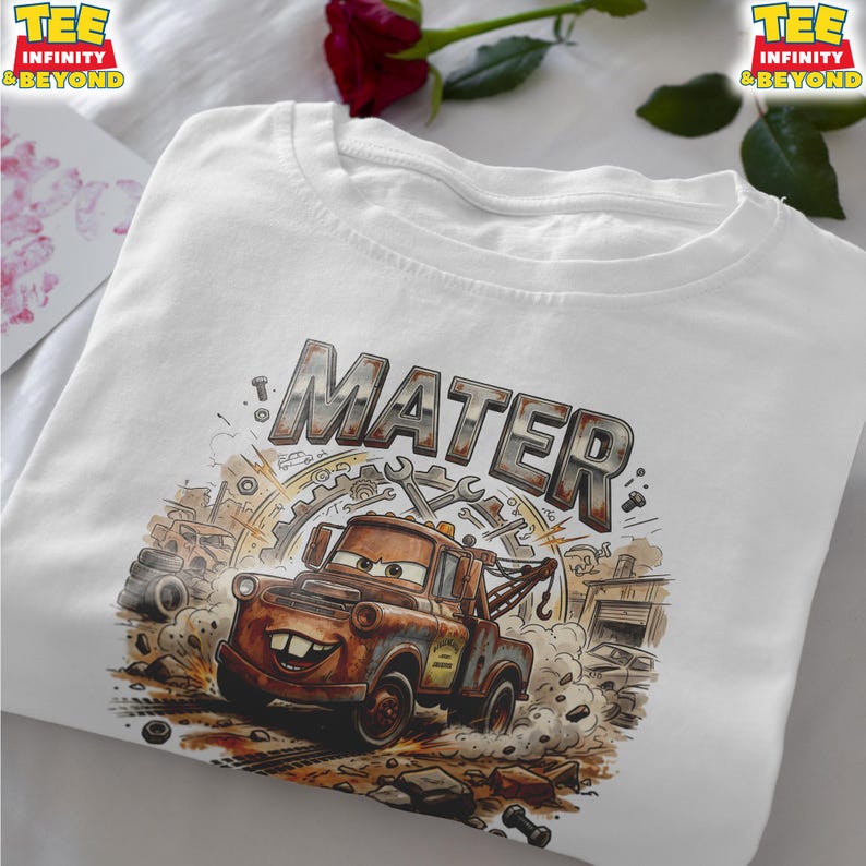May include: White t-shirt featuring a graphic of Mater from the Cars movie. The design includes the name "MATER" in a distressed metal font above an illustration of the character. The image shows Mater in a dynamic pose with a background of tools and buildings.