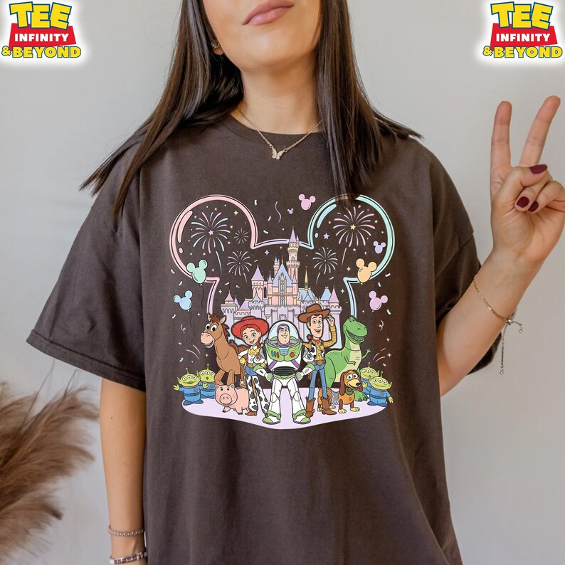 May include: Dark brown t-shirt featuring a colorful graphic of Disney's Toy Story characters in front of a castle with fireworks. The design is framed by a Mickey Mouse ear outline. The text "TEE INFINITY & BEYOND" is printed on the top right and left.