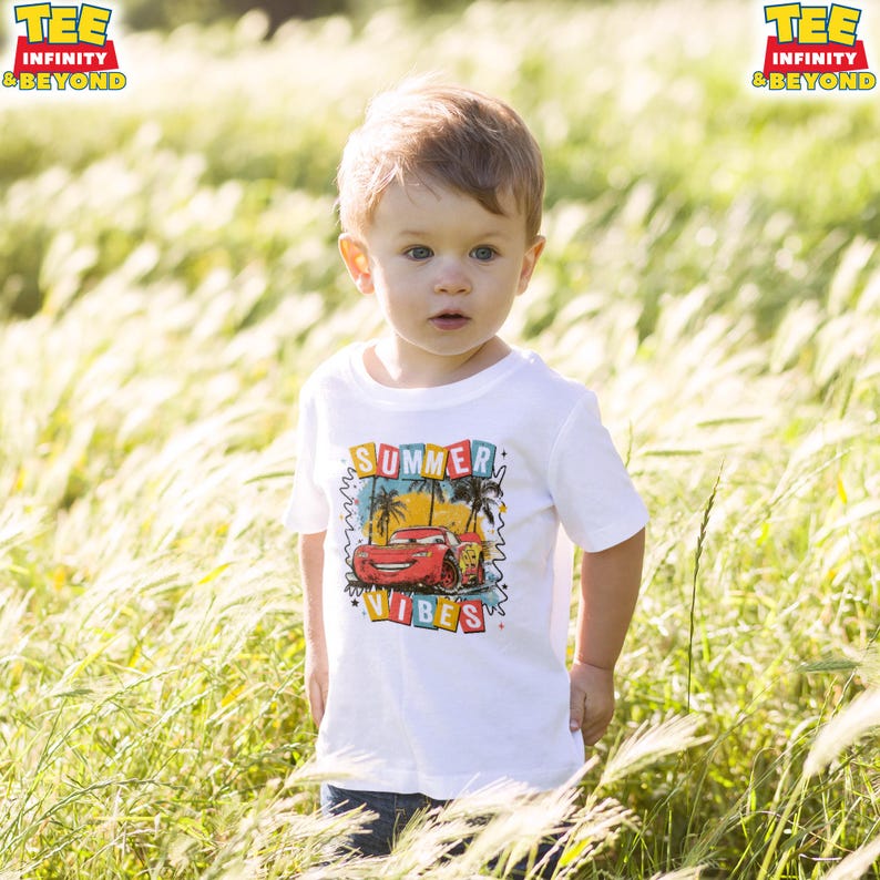 May include: A young child wearing a white t-shirt with a colorful graphic of a red race car and the words "SUMMER VIBES". The shirt has a crew neck and short sleeves. The child is standing in a field of tall grass.