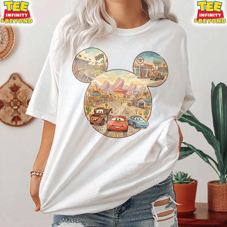 May include: White t-shirt featuring a colorful graphic of the Cars movie characters within a Mickey Mouse head outline. The design includes race tracks, desert landscapes, and Route 66 elements. The shirt has short sleeves and a relaxed fit.