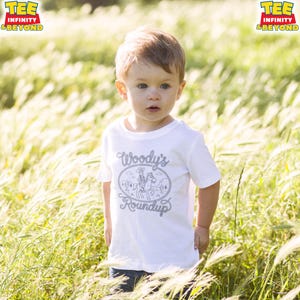 May include: A white short-sleeved t-shirt featuring a gray graphic print of a cowboy on a horse within a rope-style oval border. The text Woody's Roundup is printed in a stylized font inside the border. The shirt has a crew neckline.