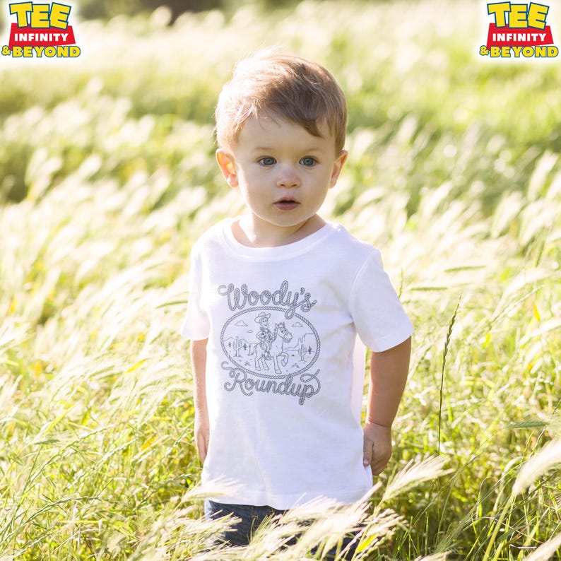 May include: A white short-sleeved t-shirt featuring a gray graphic print of a cowboy on a horse within a rope-style oval border. The text Woody's Roundup is printed in a stylized font inside the border. The shirt has a crew neckline.