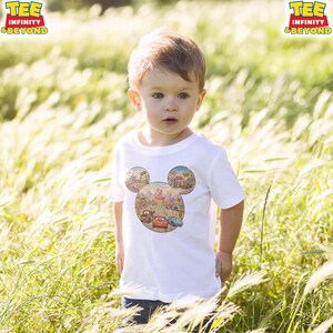 May include: A white t-shirt featuring a Mickey Mouse-shaped design with a Cars theme. The design includes illustrations of cars and buildings. The shirt is worn by a young child in a field of tall grass. The text "TEE INFINITY & BEYOND" is visible.