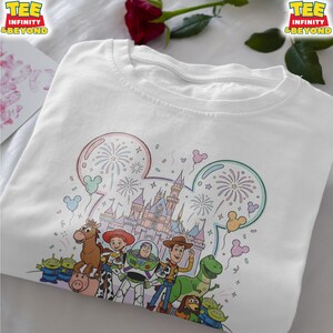 May include: White t-shirt featuring a colorful graphic of Disney characters, including Woody, Buzz Lightyear, and others, in front of a castle with fireworks. The shirt has the text "TEE INFINITY & BEYOND" at the top.