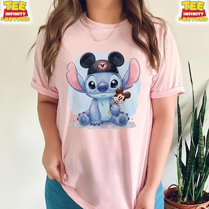 K&ouml;nnte beinhalten: Hellrosa T-Shirt mit einer Cartoon-Illustration von Stitch, der einen schwarzen Mickey Mouse-Hut tr&auml;gt und einen Mickey Mouse-Eis am Stiel h&auml;lt. Das Shirt hat hochgekrempelte &Auml;rmel und den Text "TEE INFINITY & BEYOND" oben links und rechts.