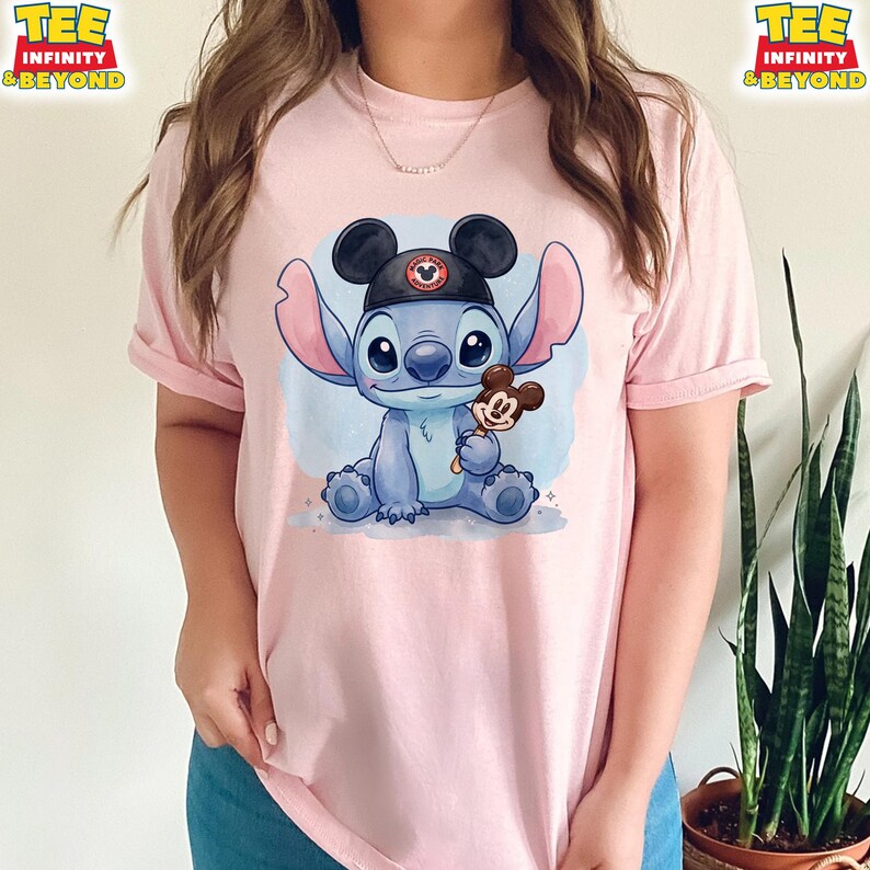 K&ouml;nnte beinhalten: Hellrosa T-Shirt mit einer Cartoon-Illustration von Stitch, der einen schwarzen Mickey Mouse-Hut tr&auml;gt und einen Mickey Mouse-Eis am Stiel h&auml;lt. Das Shirt hat hochgekrempelte &Auml;rmel und den Text "TEE INFINITY & BEYOND" oben links und rechts.