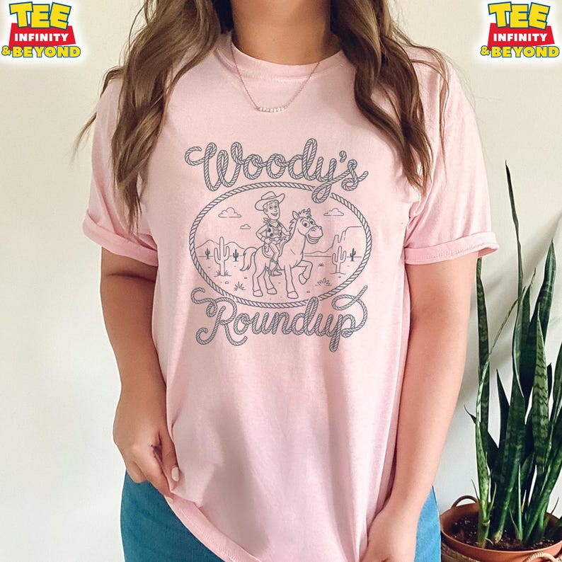 May include: A light pink short-sleeve t-shirt featuring a dark gray line art graphic of a cowboy riding a horse inside a rope-style oval frame. The text Woody's Roundup is printed in a stylized font above and below the central illustration.