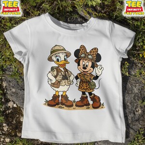 May include: White t-shirt featuring a cartoon illustration of Daisy Duck and Minnie Mouse dressed in safari outfits. Daisy Duck is wearing a khaki safari hat, vest, and boots. Minnie Mouse is wearing a leopard print bow, safari outfit, and boots. The shirt has the text "TEE INFINITY & BEYOND".
