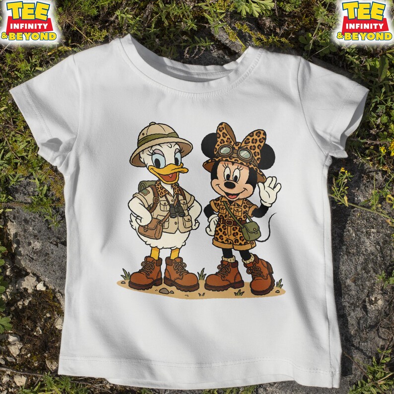 May include: White t-shirt featuring a cartoon illustration of Daisy Duck and Minnie Mouse dressed in safari outfits. Daisy Duck is wearing a khaki safari hat, vest, and boots. Minnie Mouse is wearing a leopard print bow, safari outfit, and boots. The shirt has the text "TEE INFINITY & BEYOND".