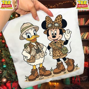 May include: White square pillow featuring cartoon characters Daisy Duck and Minnie Mouse in safari outfits. Daisy Duck is wearing a khaki safari outfit and hat, while Minnie Mouse is in a leopard print safari outfit. Both characters are wearing brown boots.