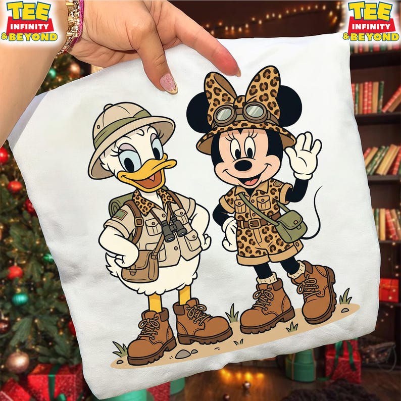 May include: White square pillow featuring cartoon characters Daisy Duck and Minnie Mouse in safari outfits. Daisy Duck is wearing a khaki safari outfit and hat, while Minnie Mouse is in a leopard print safari outfit. Both characters are wearing brown boots.