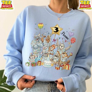 Puede incluir: Sudadera azul claro con una ilustraci&oacute;n de dibujos animados de varios personajes animales celebrando un cumplea&ntilde;os. El dise&ntilde;o incluye una estrella dorada con el n&uacute;mero 3, globos, una tarta y la palabra "Name".
