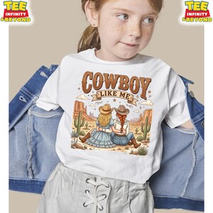 Puede incluir: Una camiseta blanca con un gr&aacute;fico de dos personas con sombreros de vaquero y ropa del oeste sentadas en un paisaje des&eacute;rtico con cactus y formaciones rocosas. El texto COWBOY LIKE ME est&aacute; impreso en marr&oacute;n sobre la ilustraci&oacute;n.