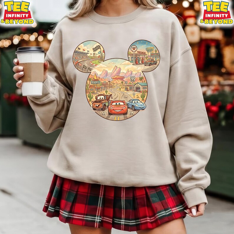 May include: Beige crewneck sweatshirt featuring a colorful graphic of Disney's Cars characters within a Mickey Mouse head shape. The design includes a race track, desert landscape, and Route 66 elements.