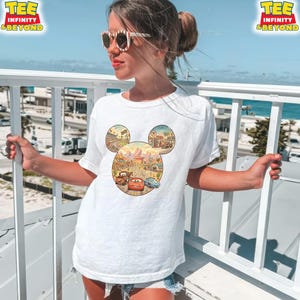 May include: White t-shirt featuring a Mickey Mouse-shaped design with a Cars movie theme. The design includes illustrations of cars, landscapes, and the text "TEE INFINITY & BEYOND". The shirt is worn by a person outdoors.