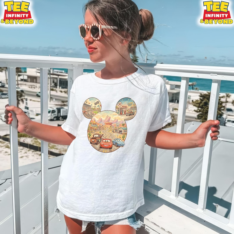 May include: White t-shirt featuring a Mickey Mouse-shaped design with a Cars movie theme. The design includes illustrations of cars, landscapes, and the text "TEE INFINITY & BEYOND". The shirt is worn by a person outdoors.