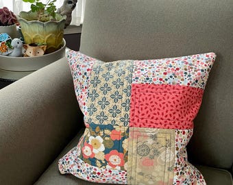 May include: Square patchwork pillow with floral and geometric patterns in shades of pink, blue, cream, and gold. The pillow is resting on a gray armchair. The pillow's design is divided into four sections.