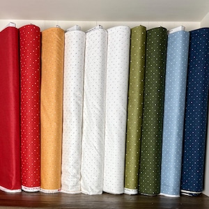 May include: A collection of fabric rolls in various colors, including red, orange, white, green, blue, and navy. Each roll is adorned with a repeating pattern of small, evenly spaced white dots. The fabrics are neatly arranged on a shelf.