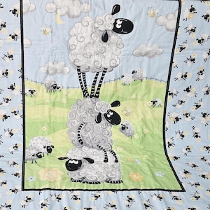May include: A baby quilt with a whimsical sheep design. The quilt features a stack of three fluffy white sheep with black faces and legs, set against a blue sky with clouds, bees, and a green field. The border is blue with sheep.