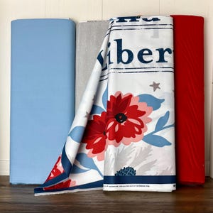 May include: A collection of fabric rolls in various colors. A light blue, a gray with white dots, and a white fabric with red flowers and the word "Liberty" printed on it. A solid red fabric roll is also visible.