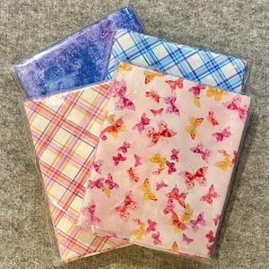 May include: Four fabric bundles in clear plastic packaging. The fabrics feature various patterns, including a blue watercolor design, a blue and white plaid, a pink and yellow plaid, and a pink fabric with butterfly illustrations.