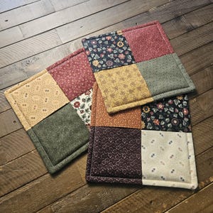May include: Three quilted fabric pot holders with a patchwork design. The pot holders are made of different floral patterned fabrics in shades of red, green, yellow, and brown.