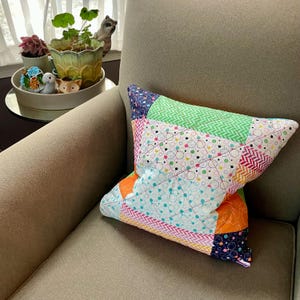 May include: A colorful patchwork pillow with geometric patterns in shades of green, pink, blue, orange, and white. The pillow is sitting on a beige armchair.