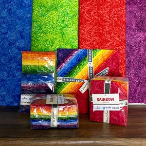May include: A collection of colorful fabric bundles, including rainbow-themed packs and patterned fabrics in red, green, blue, and purple. The bundles are wrapped and labeled with the "Island Batik" brand.